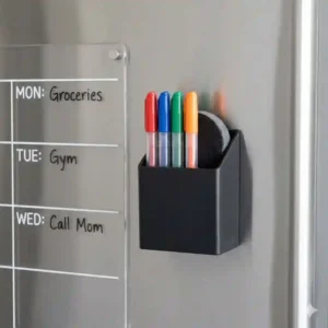 Small magnetic dry erase marker holder holding fine-tip pens next to a weekly refrigerator calendar.